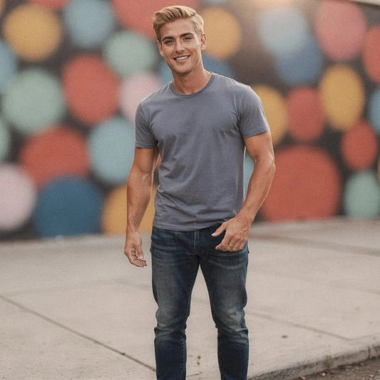 Man wearing blue jeans and a gray t shirt stands outdoors, colorful bokeh background, smiling