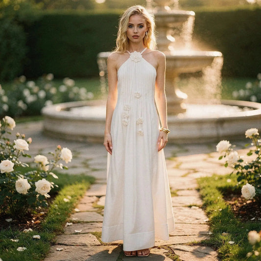 Woman wearing a white halter maxi dress with floral accents, standing on a garden path near a fountain